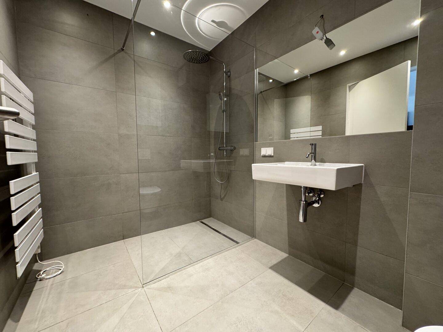 Modernes Badezimmer mit grauen Fliesen, ebenerdiger Dusche und beheiztem Handtuchhalter.