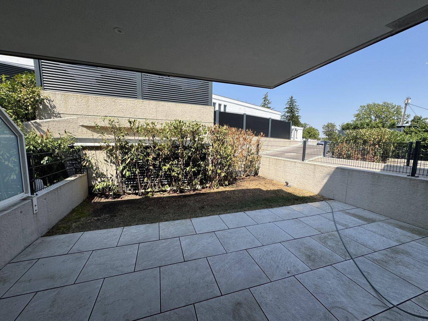 Überdachte Terrasse mit großen Fliesen und kleinem Gartenbereich, umgeben von Hecken.