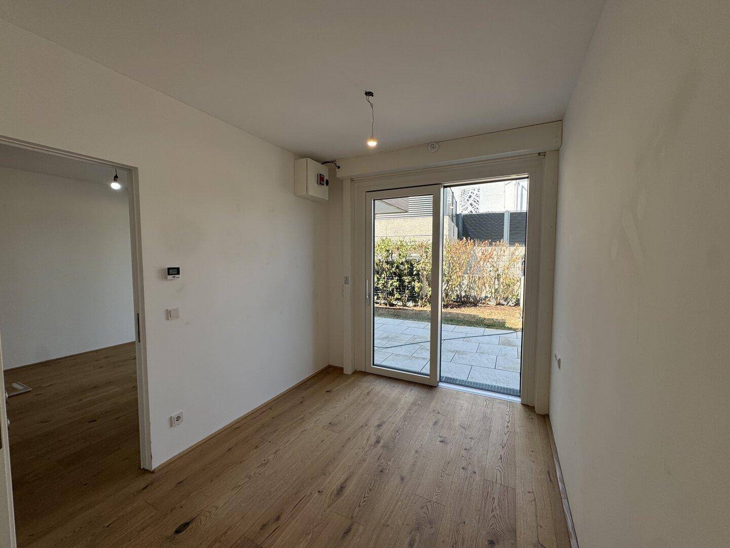 Helles Zimmer mit Holzboden und großer Schiebetür, die zu einem begrünten Außenbereich führt.