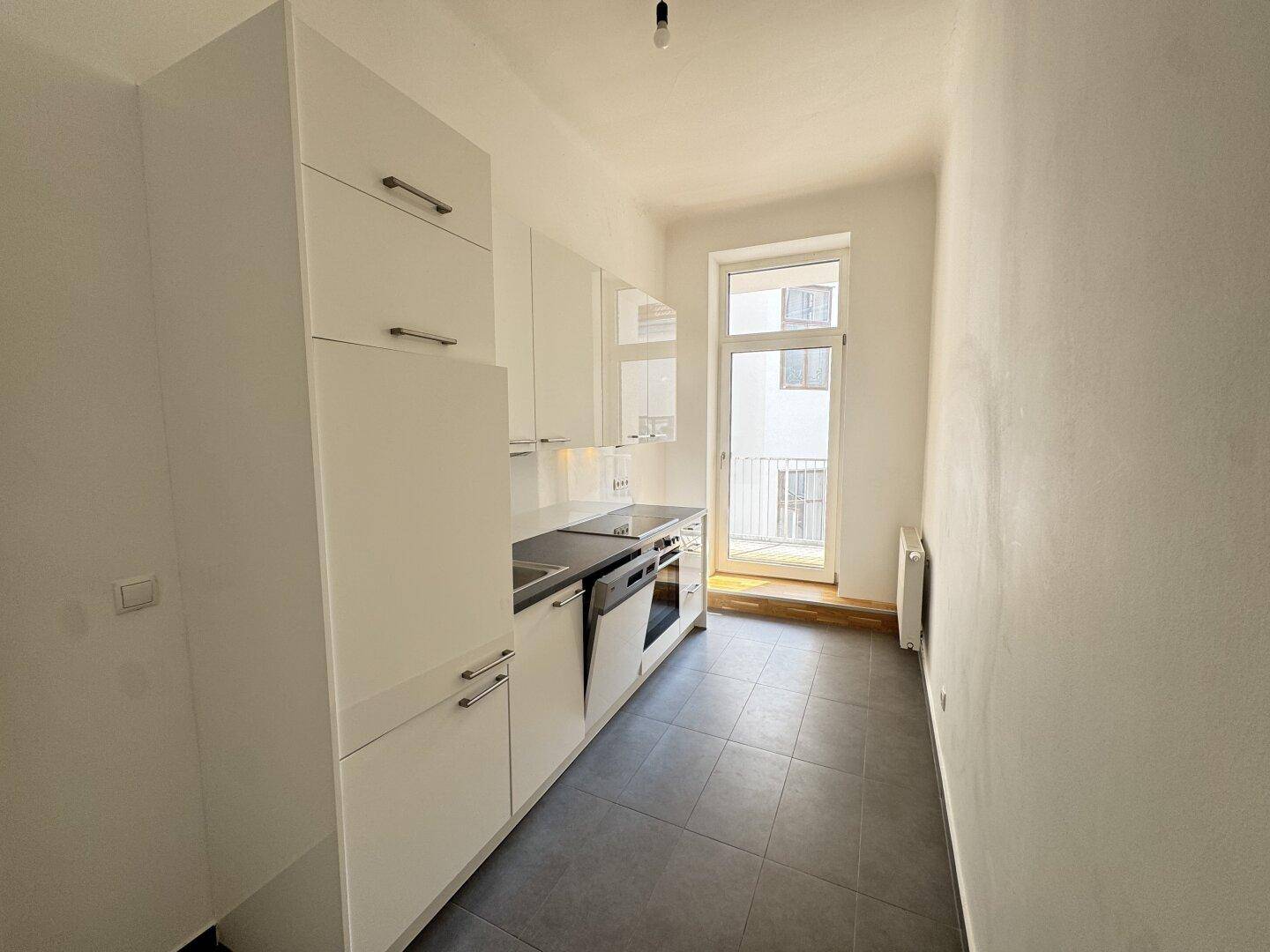 Schmale, moderne Küche mit weißen Schränken, Einbaugeräten und Zugang zum Balkon.