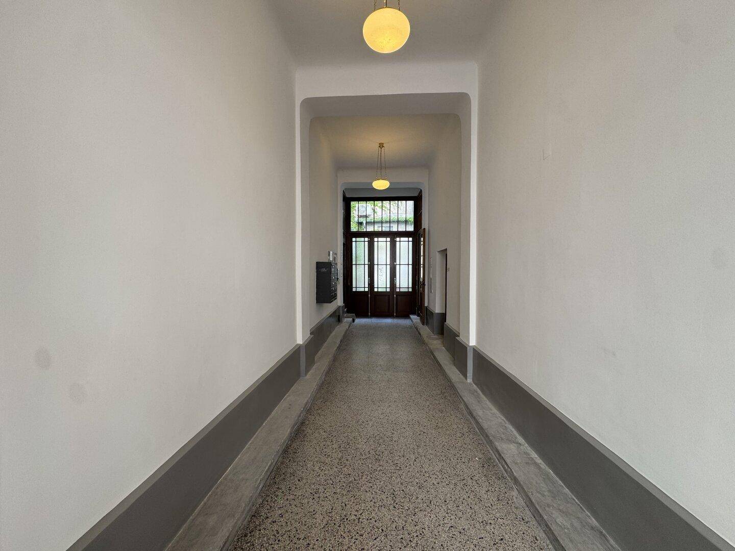 Langer, heller Flur mit Terrazzoboden und doppelter Glastür am Ende des Ganges.