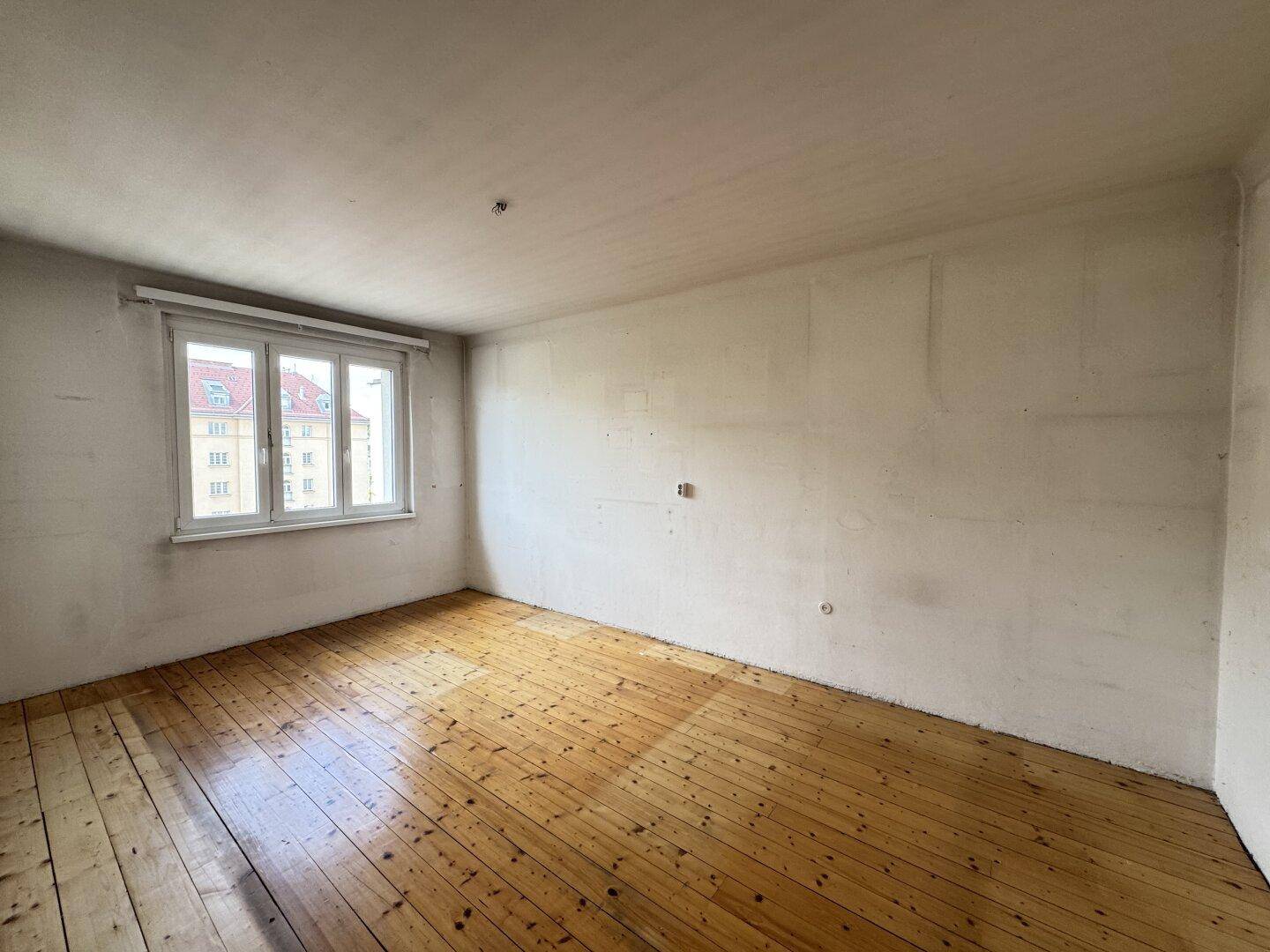 Geräumiger, leerer Raum mit Holzdielenboden und einem großen Fenster, das viel Licht hereinlässt.