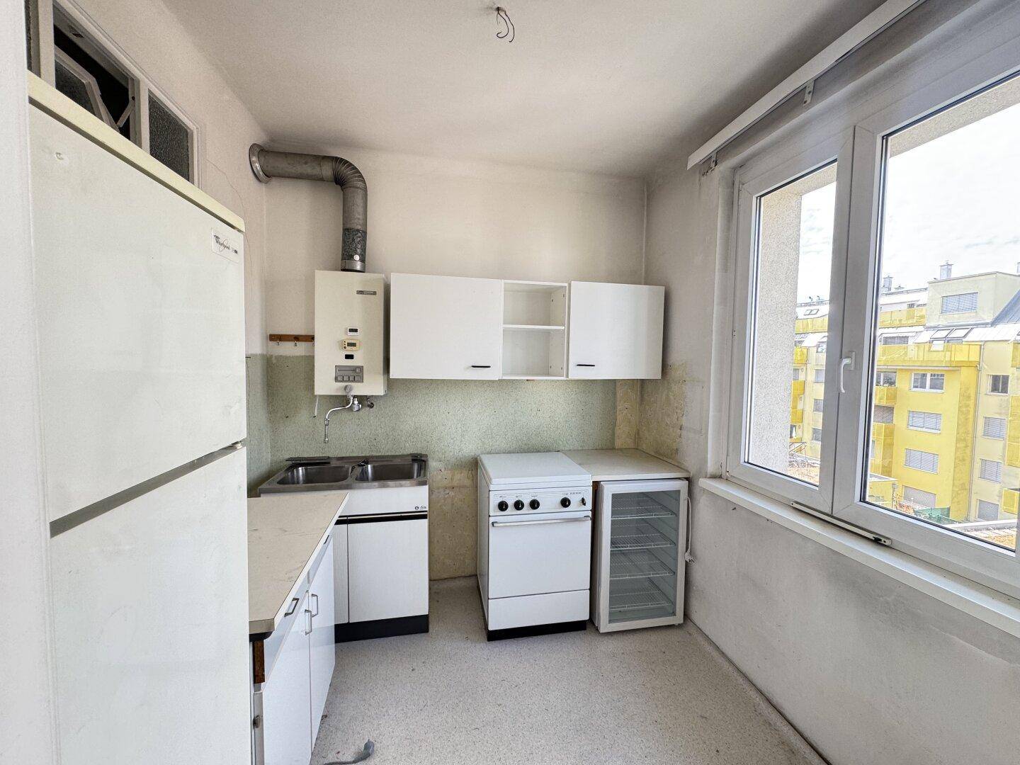 Kleine Küche mit älteren Geräten, Spüle und Hängeschränken, hell durch großes Fenster.