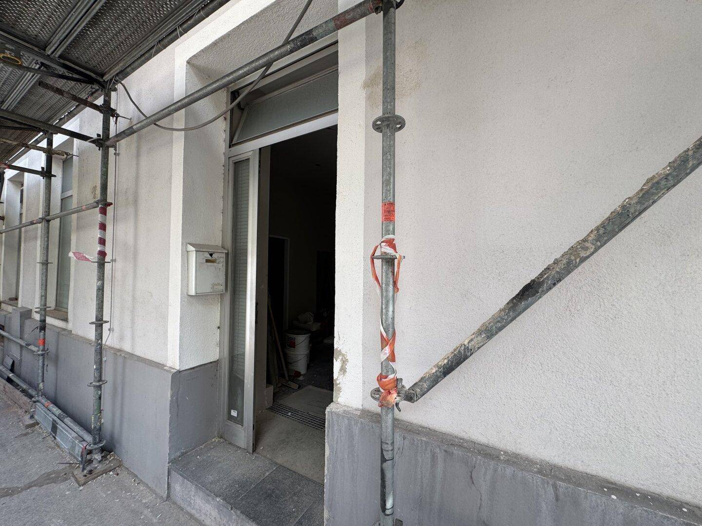 Eingangsbereich eines Gebäudes unter Baugerüst, mit einer einfachen Tür und einem Briefkasten an der Wand.