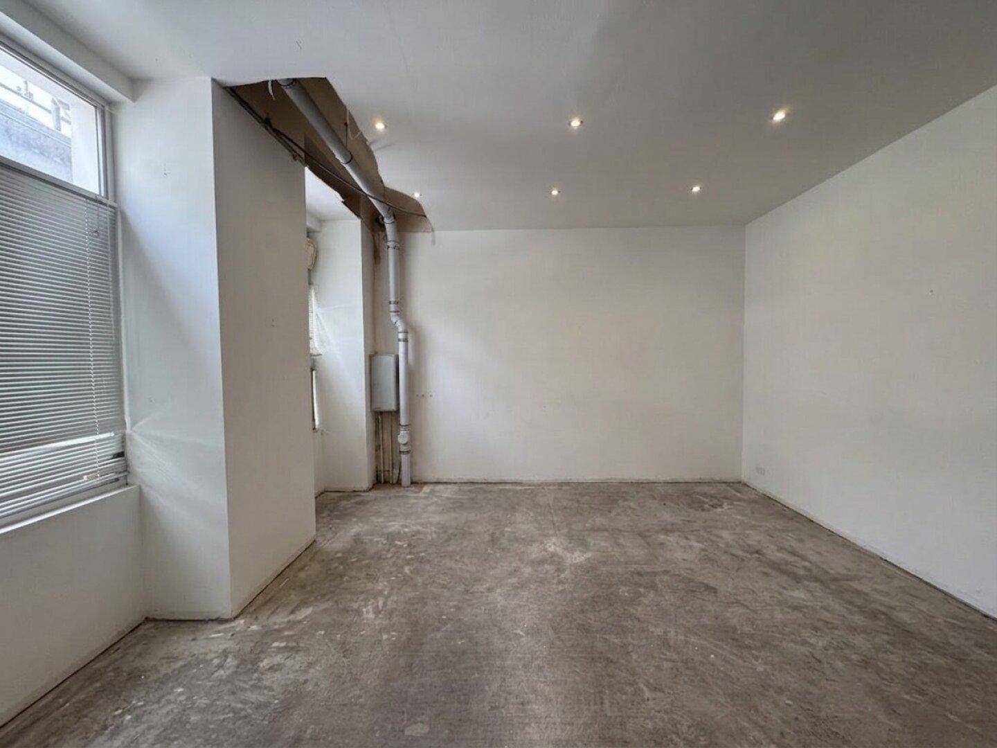 Leerer Raum mit Betonboden und weißen Wänden, sichtbaren Rohren und einem Fenster mit Jalousien.