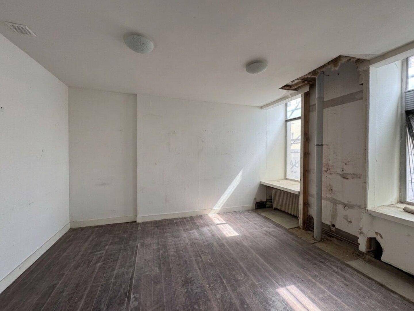 Unfertiger Innenraum mit dunklem Holzfußboden, weißen Wänden und sichtbaren Renovierungsspuren an den Fenstern.