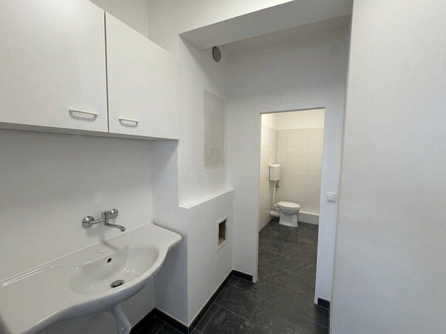 Kleiner Waschraum mit Waschbecken und weißen Hängeschränken, angrenzend an eine separate Toilette mit dunklen Fliesen.