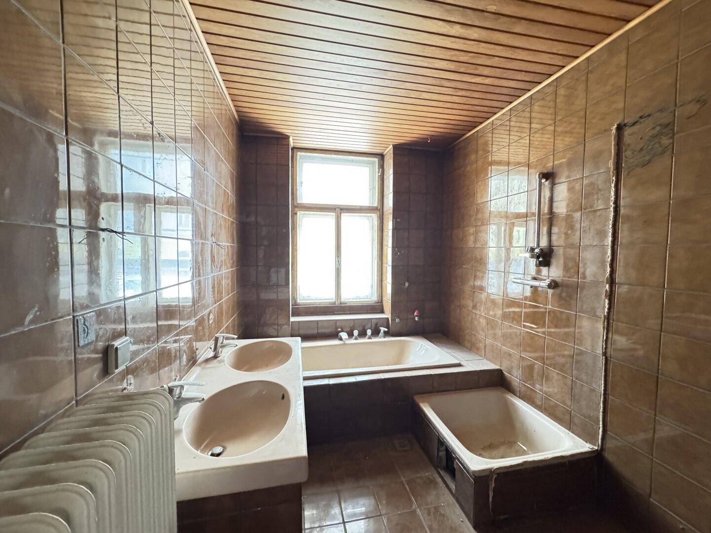 Badezimmer im 80er-Jahre-Stil mit braunen Fliesen, Doppelwaschbecken und Badewanne, renovierungsbedürftig.