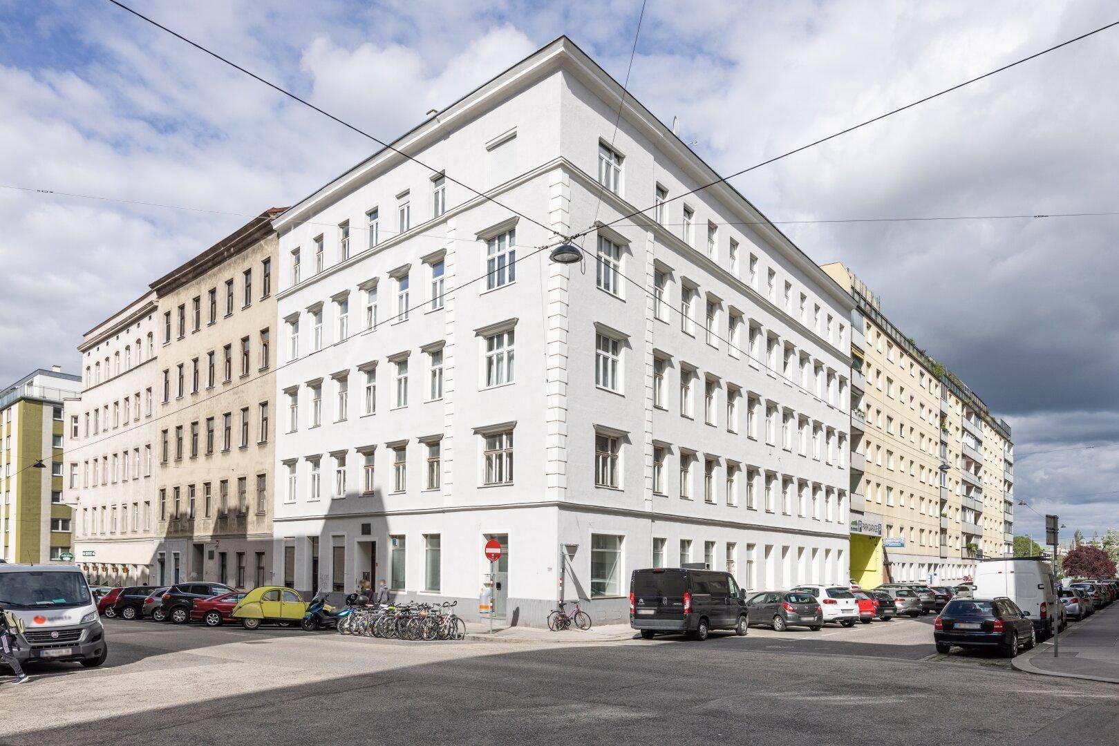 Klassisches Eckgebäude in städtischer Umgebung mit vielen Fenstern und geparkten Autos auf der Straße.