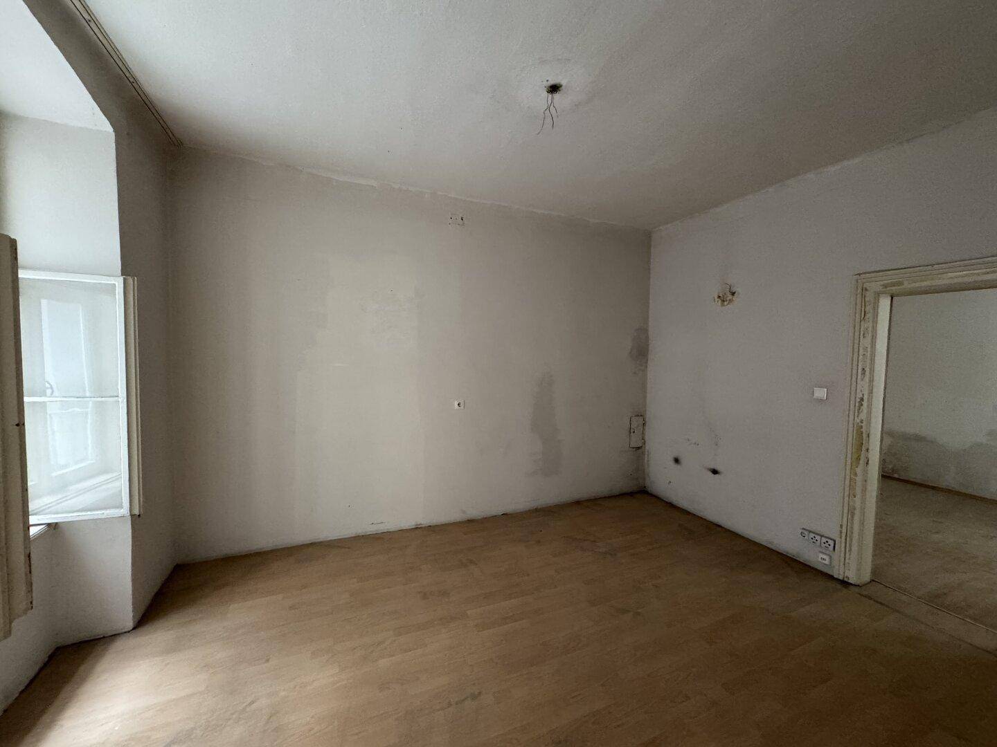Geräumiger, unrenovierter Raum mit Holzboden und Zugang zu einem weiteren Zimmer.