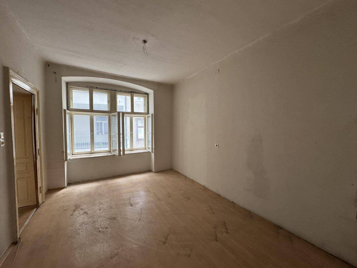 Unrenovierter Raum mit großen Fenstern und abgenutztem Holzboden, bereit zur Sanierung.
