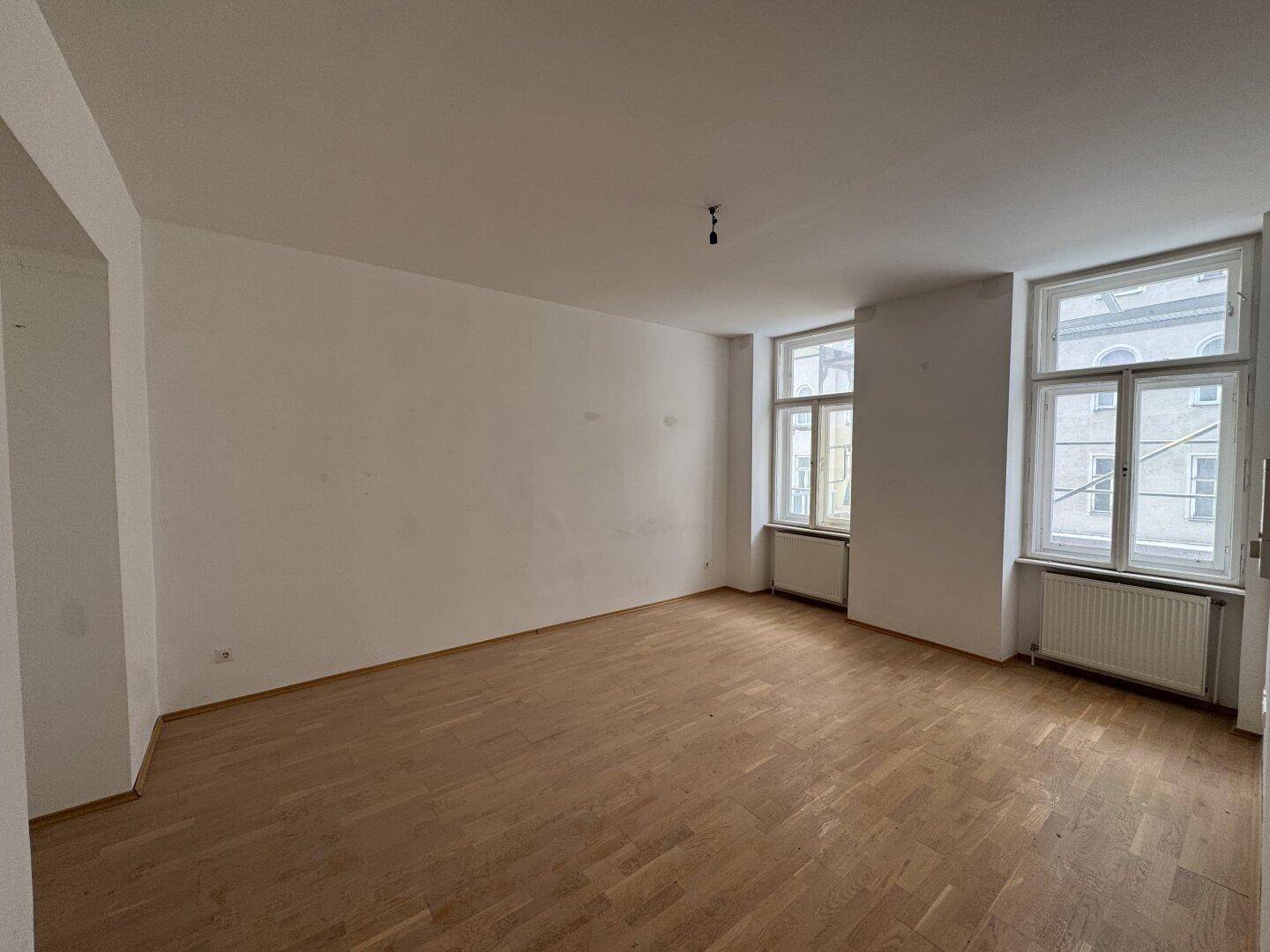 Geräumiger, leerer Raum mit Holzboden und zwei großen Fenstern.