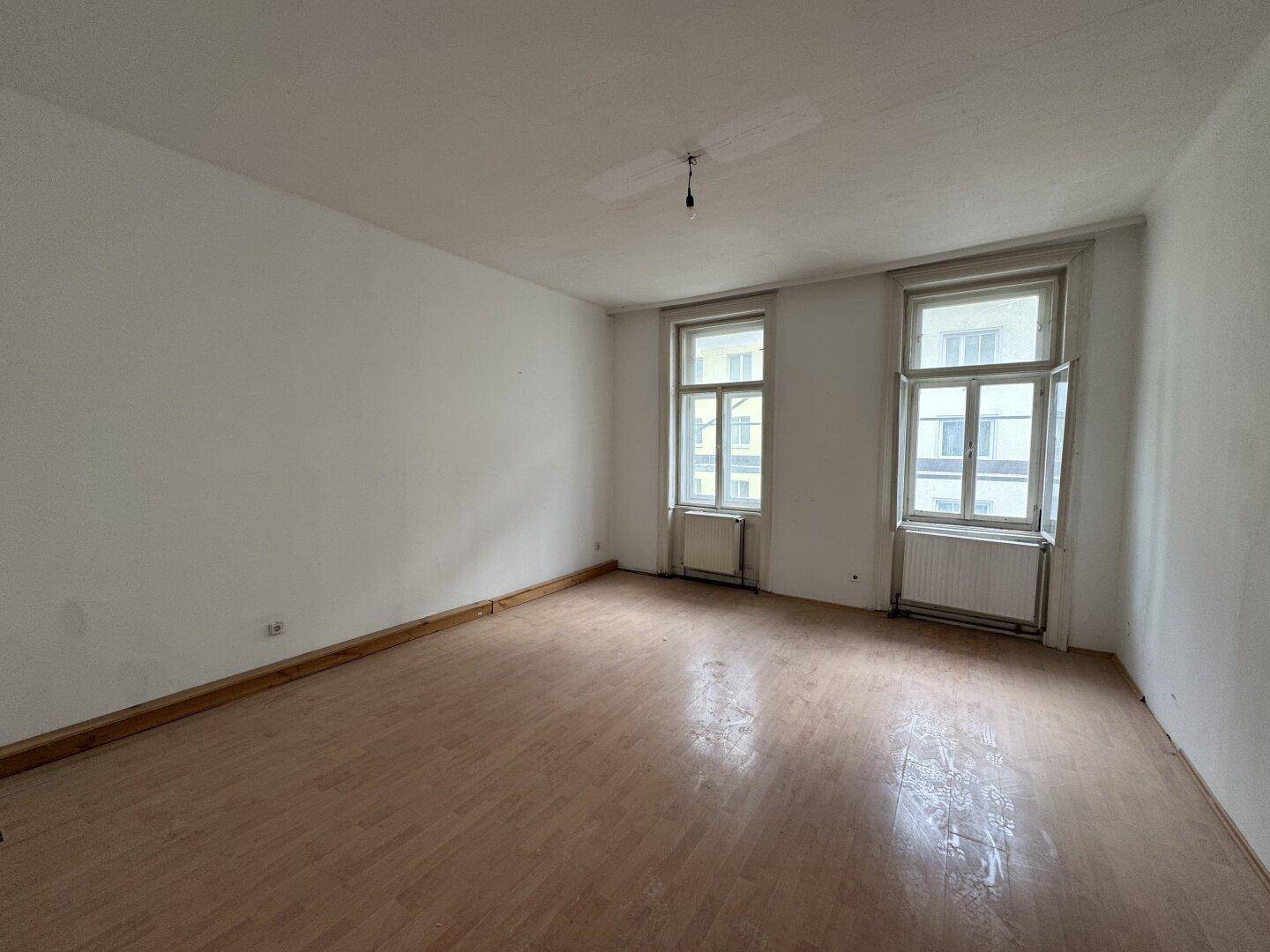Leerer Raum mit Laminatboden, hohen Fenstern und Heizkörpern, bereit für die Renovierung.