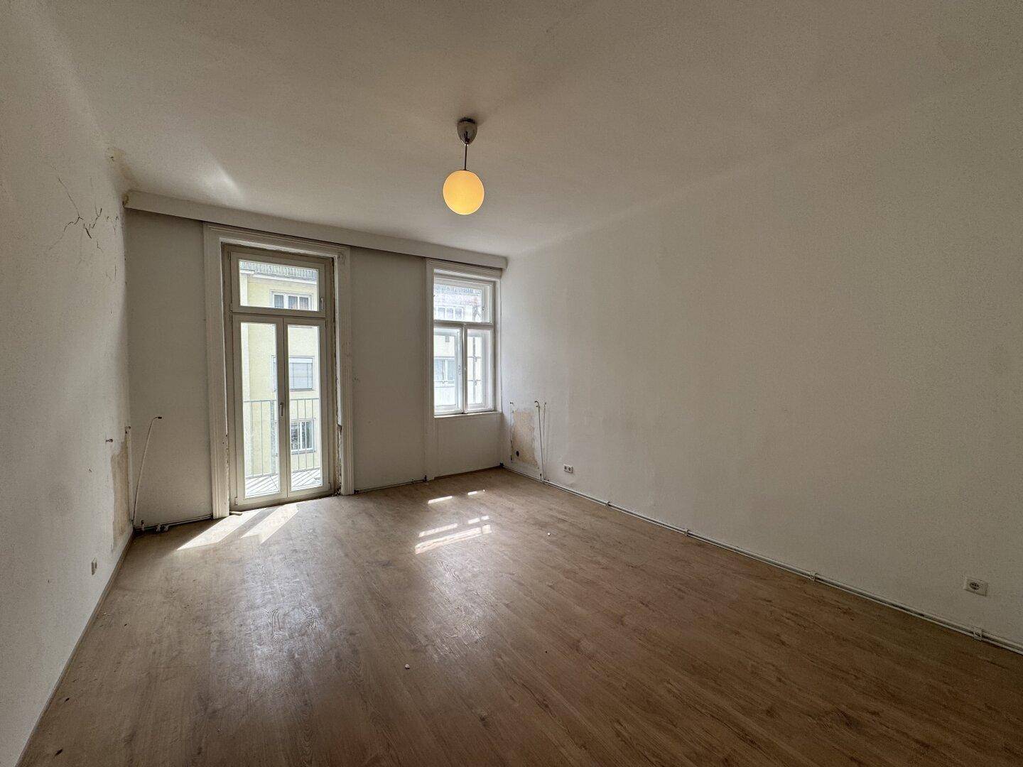 Helles, leeres Zimmer mit Holzboden und zwei Fenstern, eines davon mit französischem Balkon.