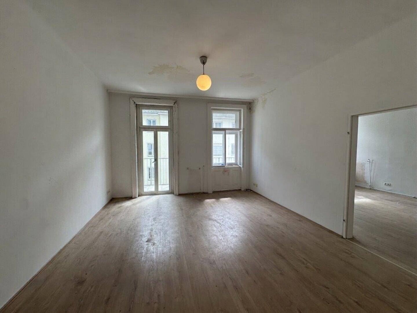 Geräumiges leeres Zimmer mit Holzboden, zwei Fenstern und Zugang zu einem französischen Balkon.
