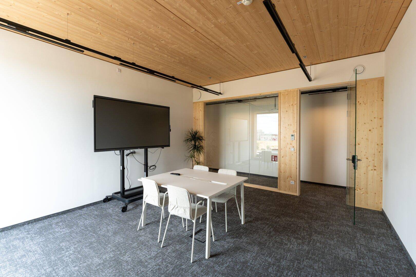 Moderner Konferenzraum mit Holzdecke, Teppichboden, großem Bildschirm und Glastrennwänden.