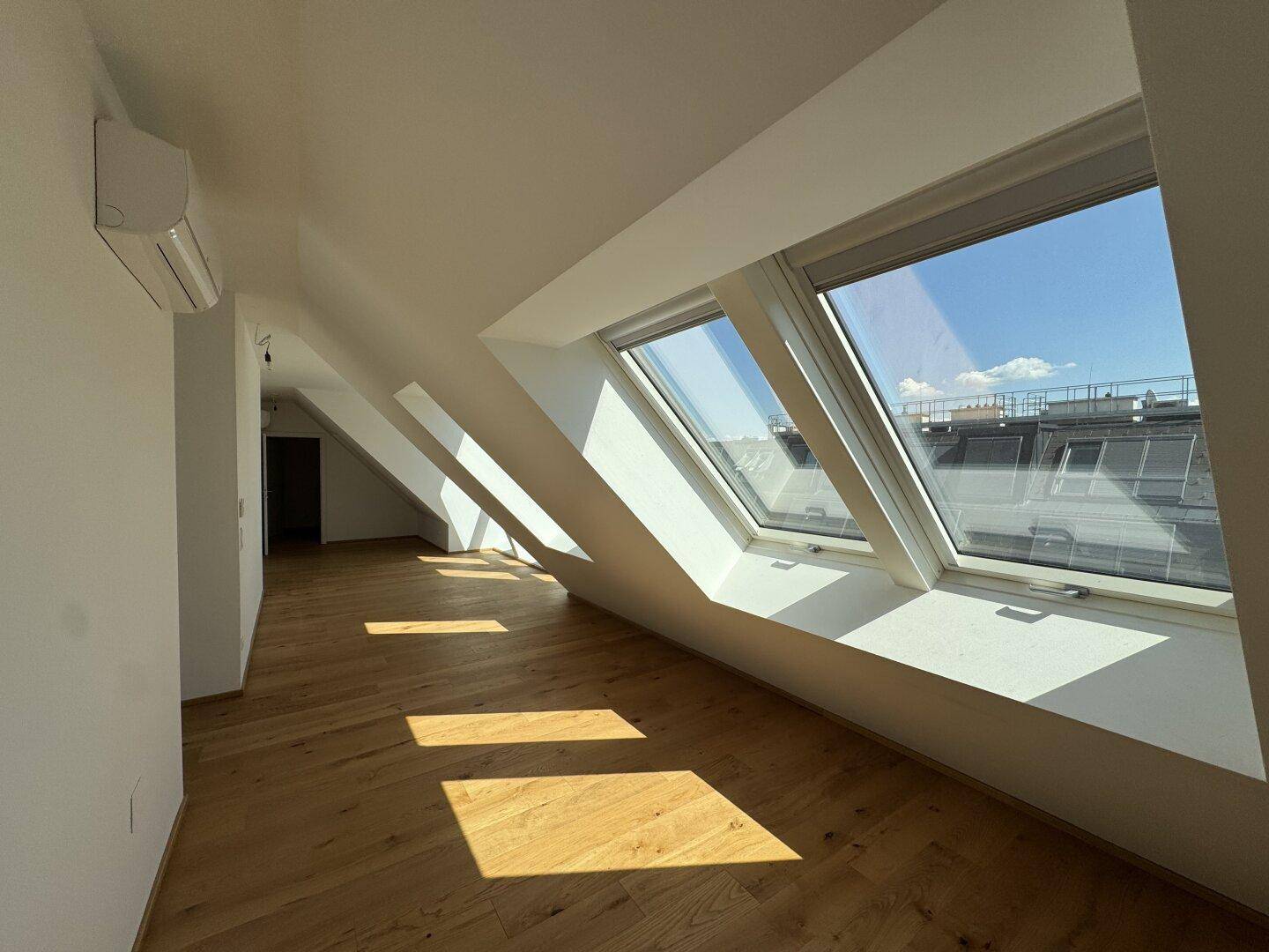 Lichtdurchfluteter Raum mit Holzboden, Dachschrägen, mehreren Dachfenstern und Klimaanlage.