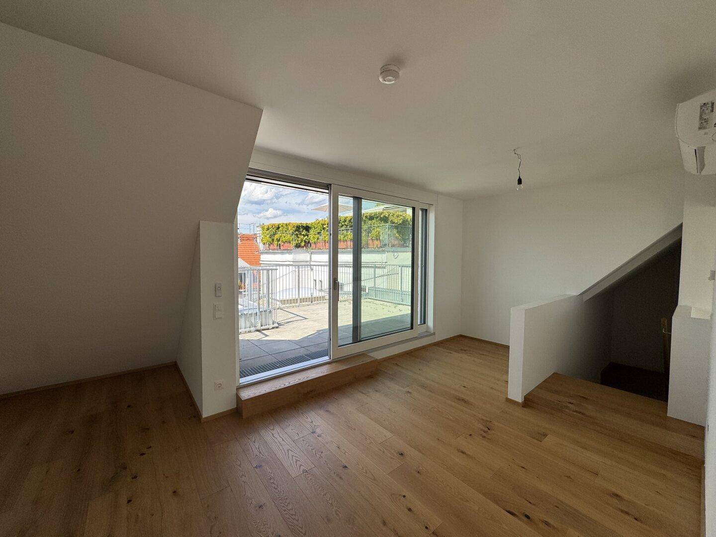Heller Wohnbereich mit Holzboden, Dachschräge, Terrassenzugang und offener Treppe zum unteren Geschoss.