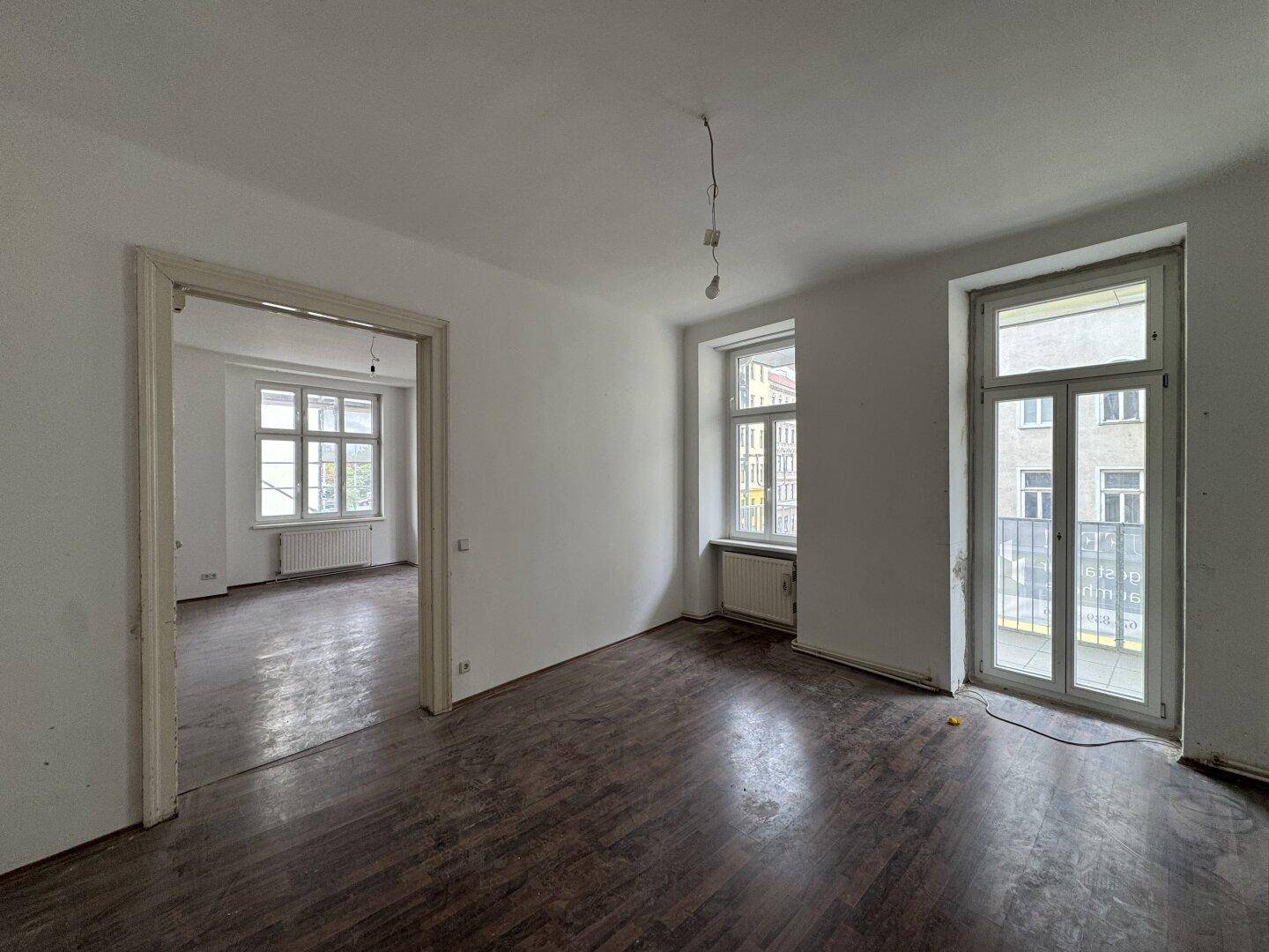 Geräumiger, heller Raum mit dunklem Parkettboden, Zugang zu einem weiteren Zimmer und französischem Balkon.
