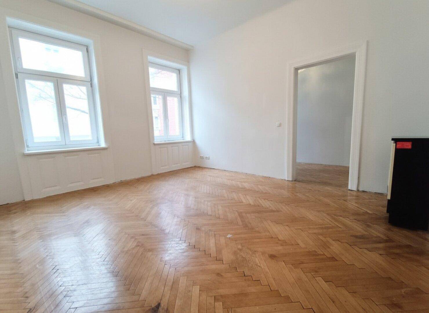 Helles, leeres Zimmer mit klassischem Fischgrätparkett und zwei großen Fenstern.