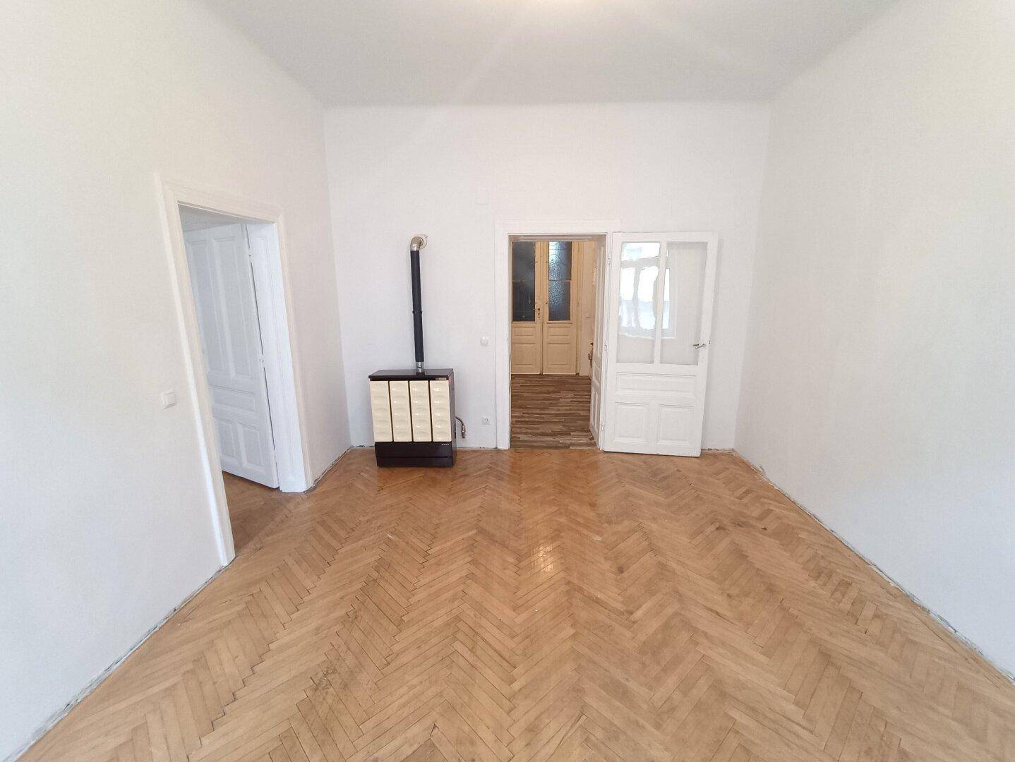 Geräumiges Zimmer mit Fischgrätparkett und einem traditionellen Ofen als Heizquelle.
