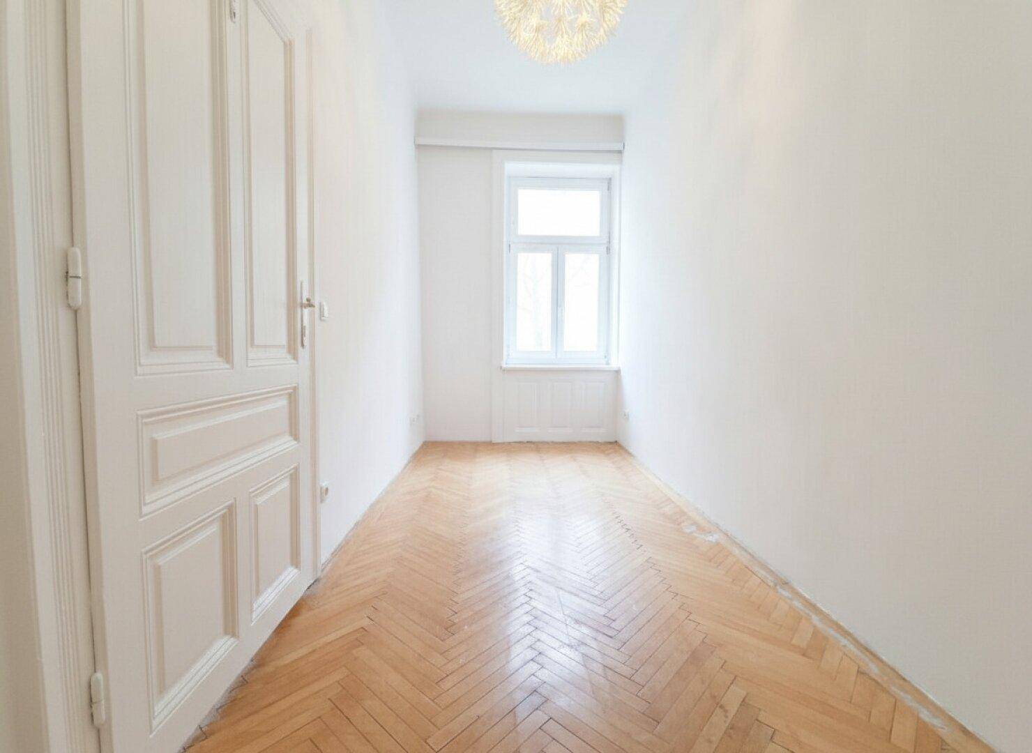 Schmales Zimmer mit Fischgrätparkett, einem Fenster und weiß gestrichenen Wänden.