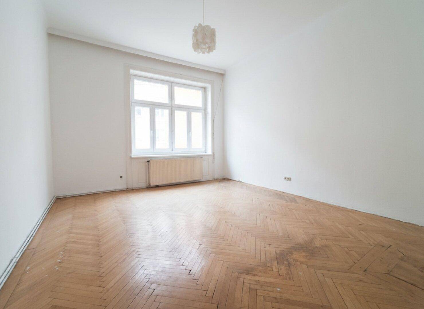 Helles Zimmer mit Fischgrätparkett, einem großen Fenster und einem Heizkörper darunter.