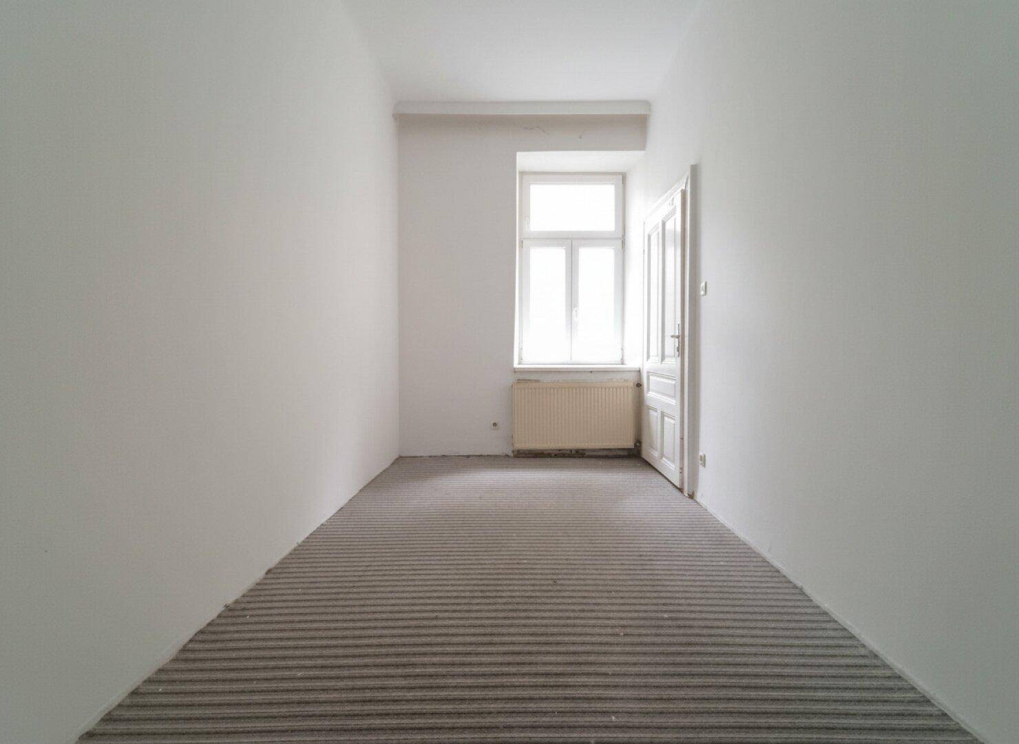 Schmales Zimmer mit gestreiftem Teppichboden, einem Fenster und einem Heizkörper.