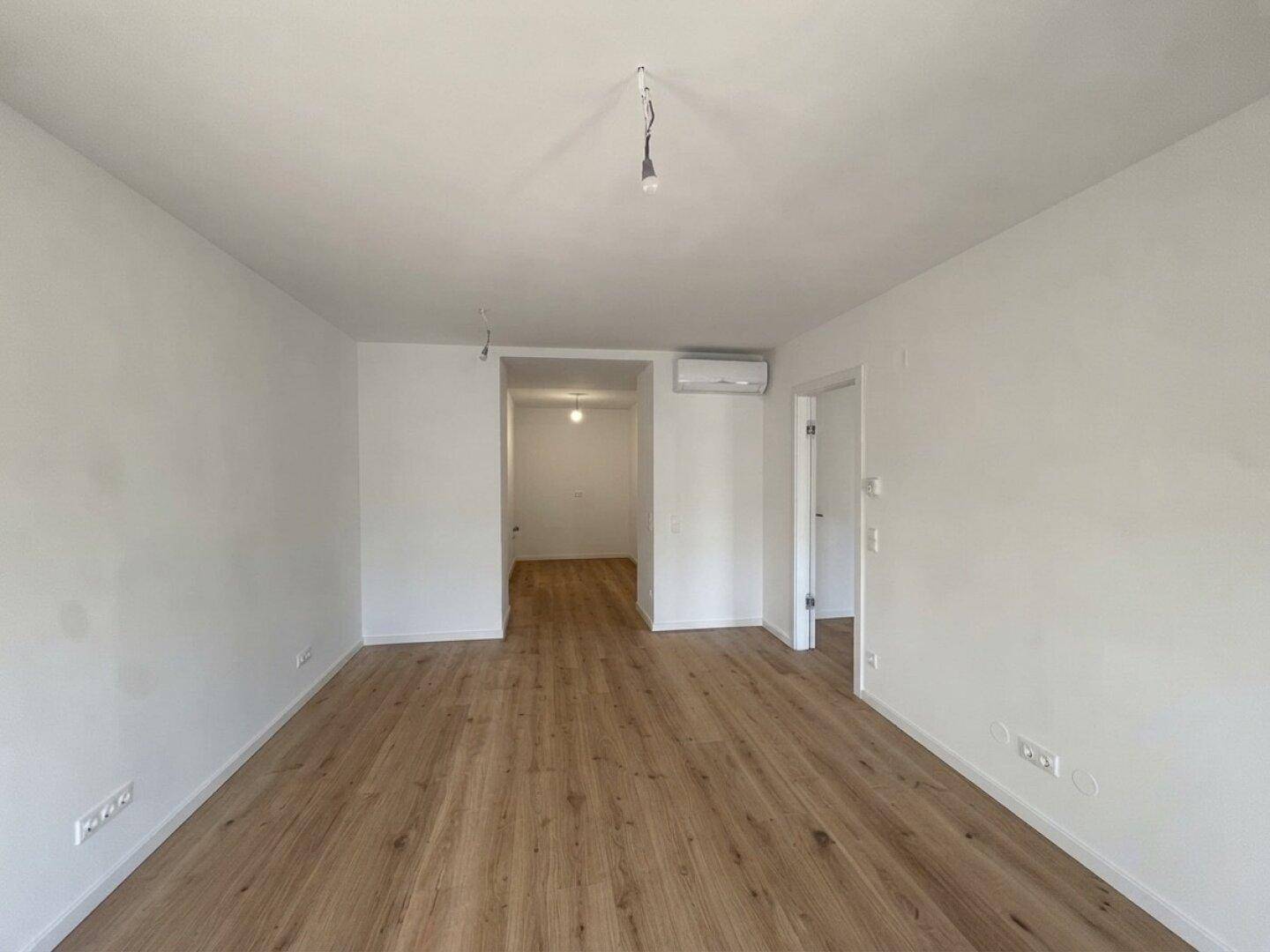Heller, leerer Wohnbereich mit Holzboden und Zugang zu weiteren Räumen.