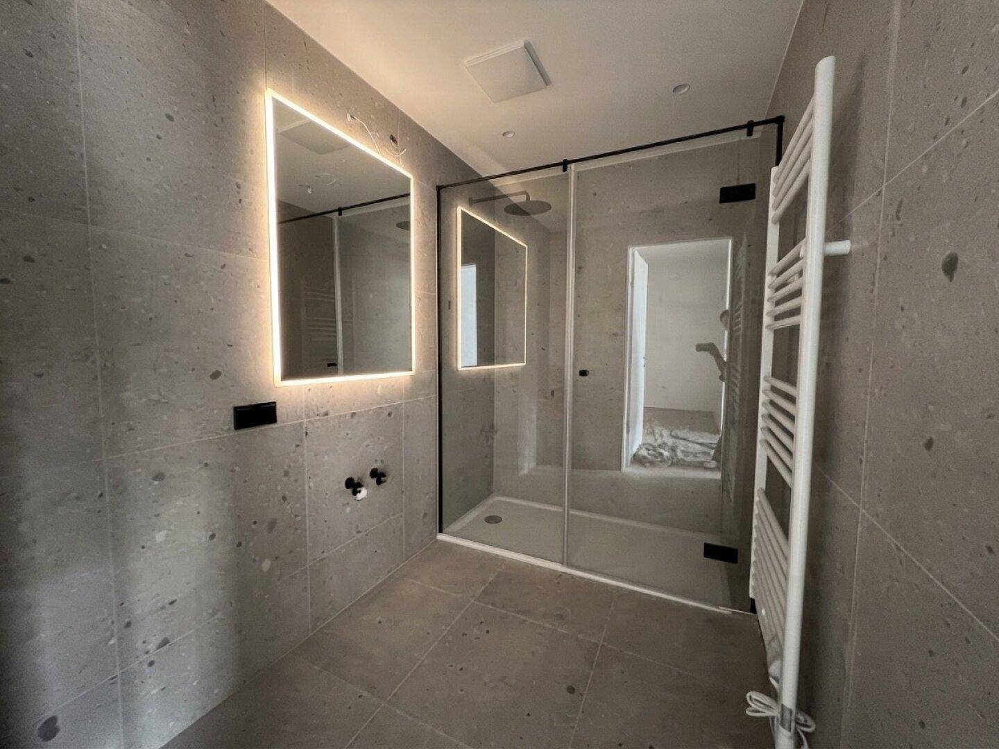 Modernes Badezimmer mit grauen Fliesen, begehbarer Dusche und beleuchtetem Spiegel.