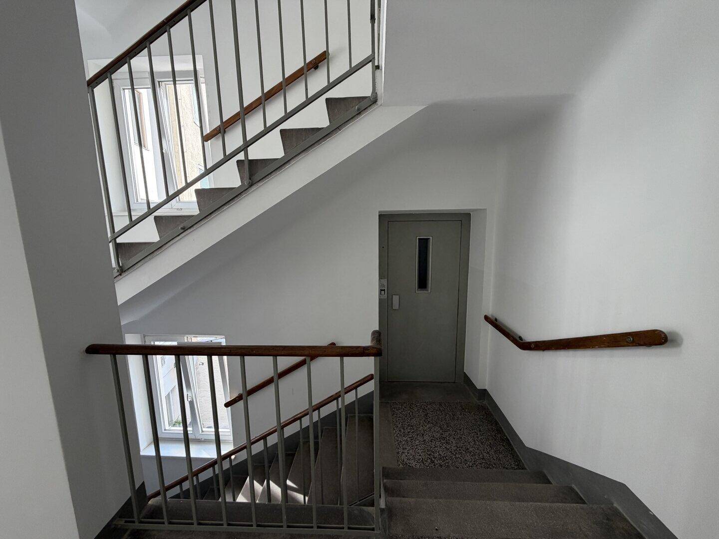 Treppenhaus mit Metallgeländer, Holzhandlauf und Zugang zu einem Aufzug.