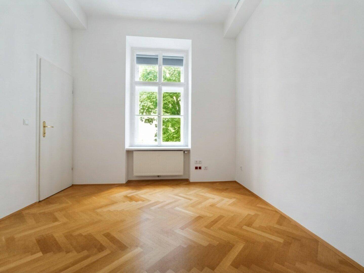Leerer Raum mit Fischgrätparkett und einem großen Fenster, das einen schönen Blick ins Grüne bietet.