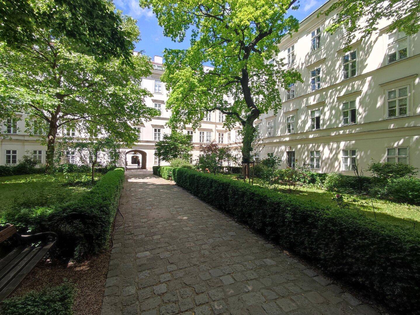 Grüner Innenhof mit gepflastertem Weg, alten Bäumen und einer Sitzbank zum Entspannen.