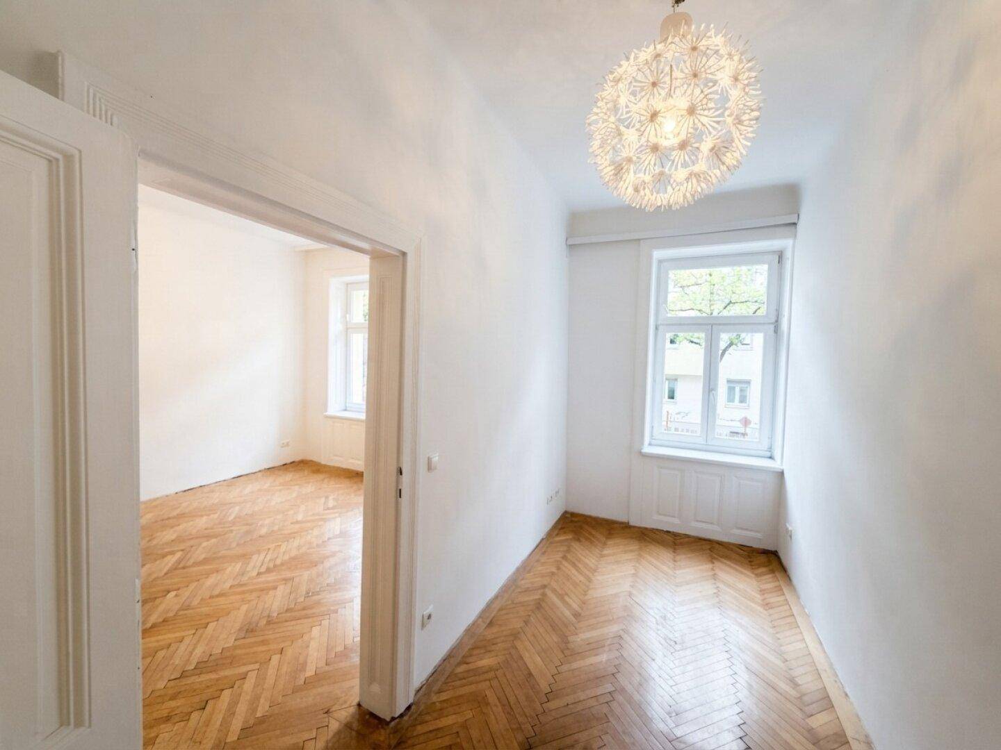 Leeres Zimmer mit Fischgrätparkett und einem Fenster, das viel Tageslicht hereinlässt.
