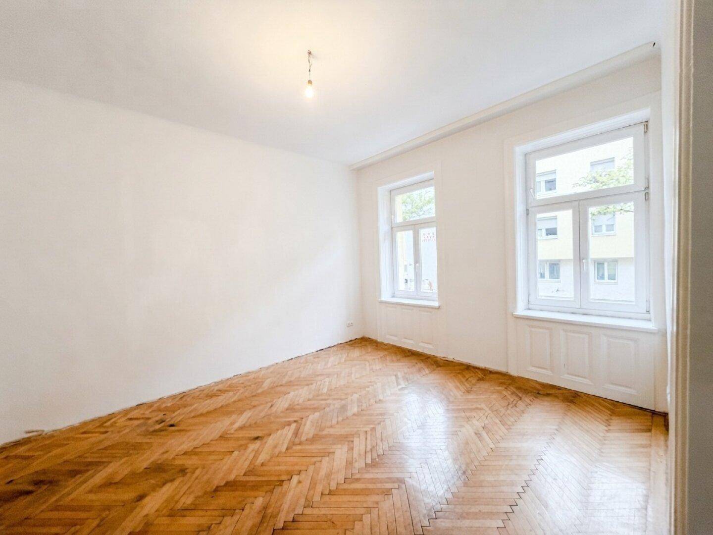 Geräumiges, leeres Zimmer mit zwei großen Fenstern und klassischem Fischgrätparkettboden.