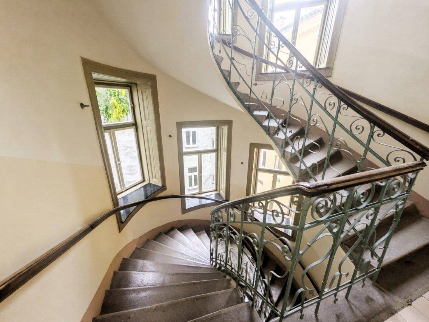 Historisches Treppenhaus mit schmiedeeisernem Geländer und großen Fenstern, die viel Licht spenden.
