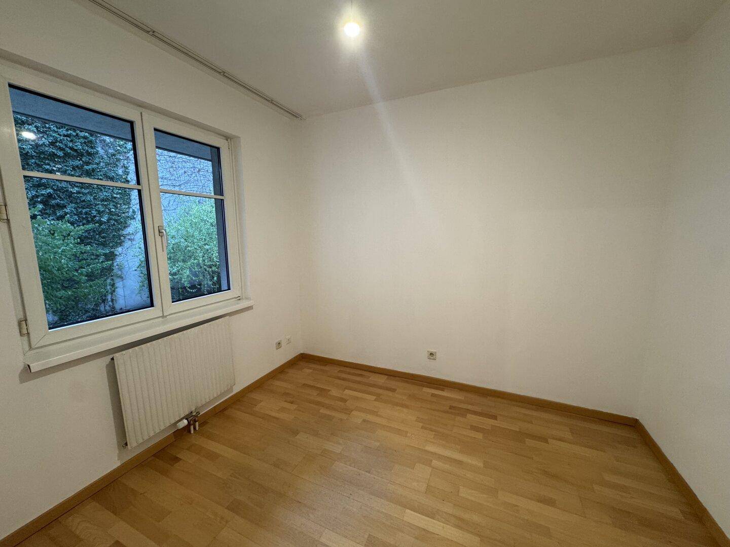 Helles Schlafzimmer mit Parkettboden und einem Fenster, das den Blick ins Grüne freigibt.