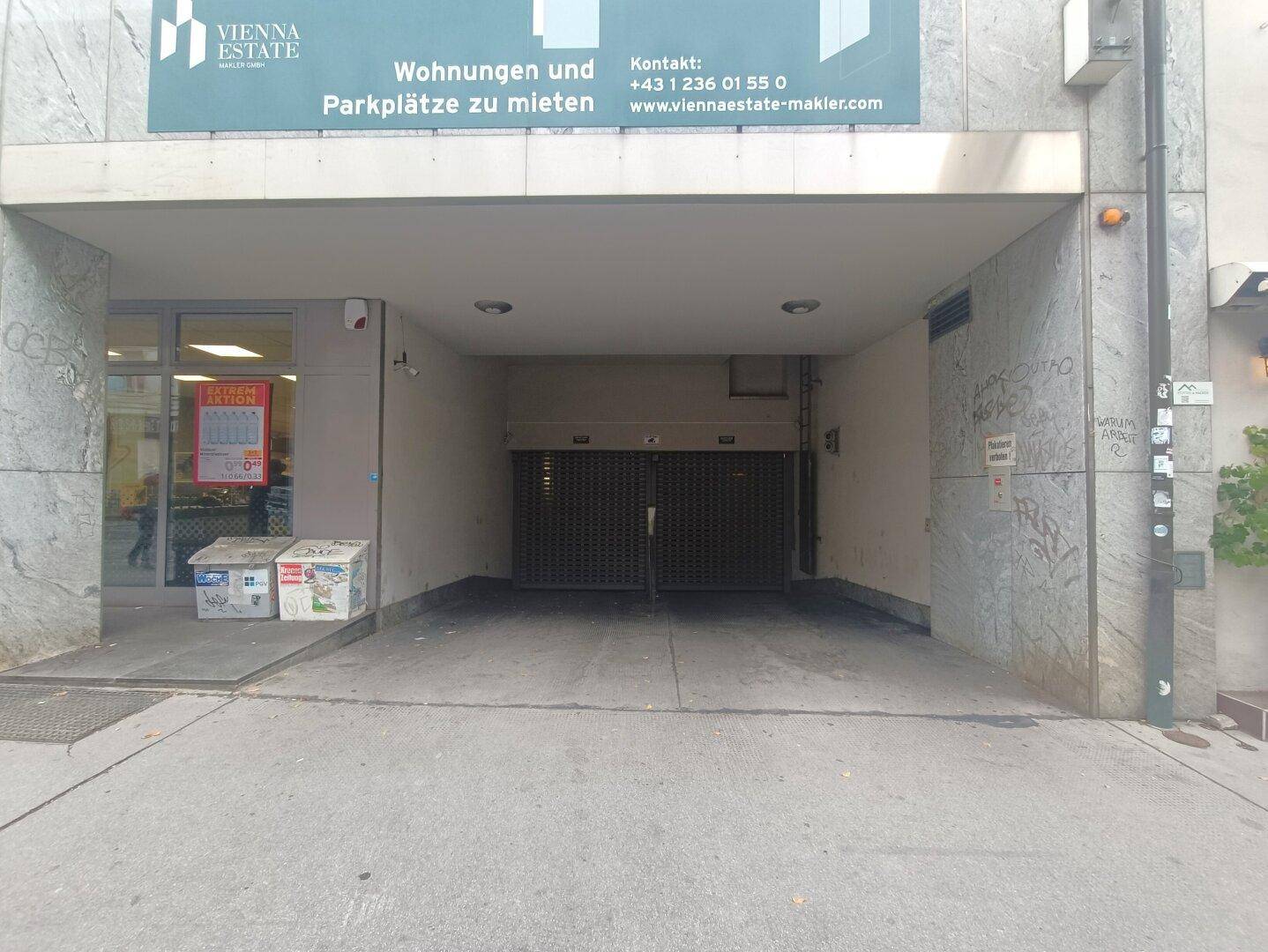 Eingang zu einer Tiefgarage von der Straße aus, mit Rolltoren und einer Werbetafel darüber.