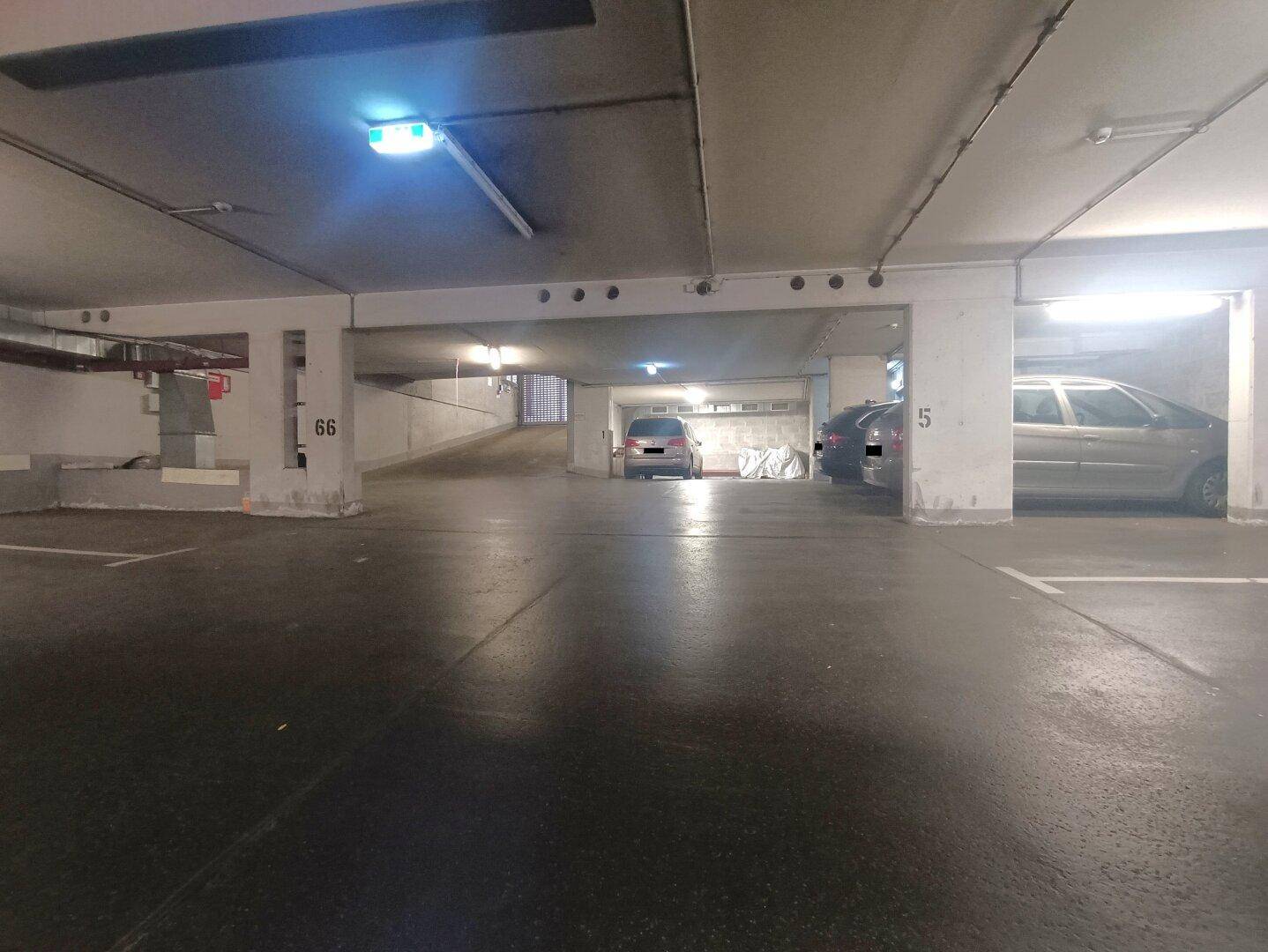 Weitläufige Tiefgarage mit geparkten Autos, einer Rampe im Hintergrund und heller Beleuchtung.