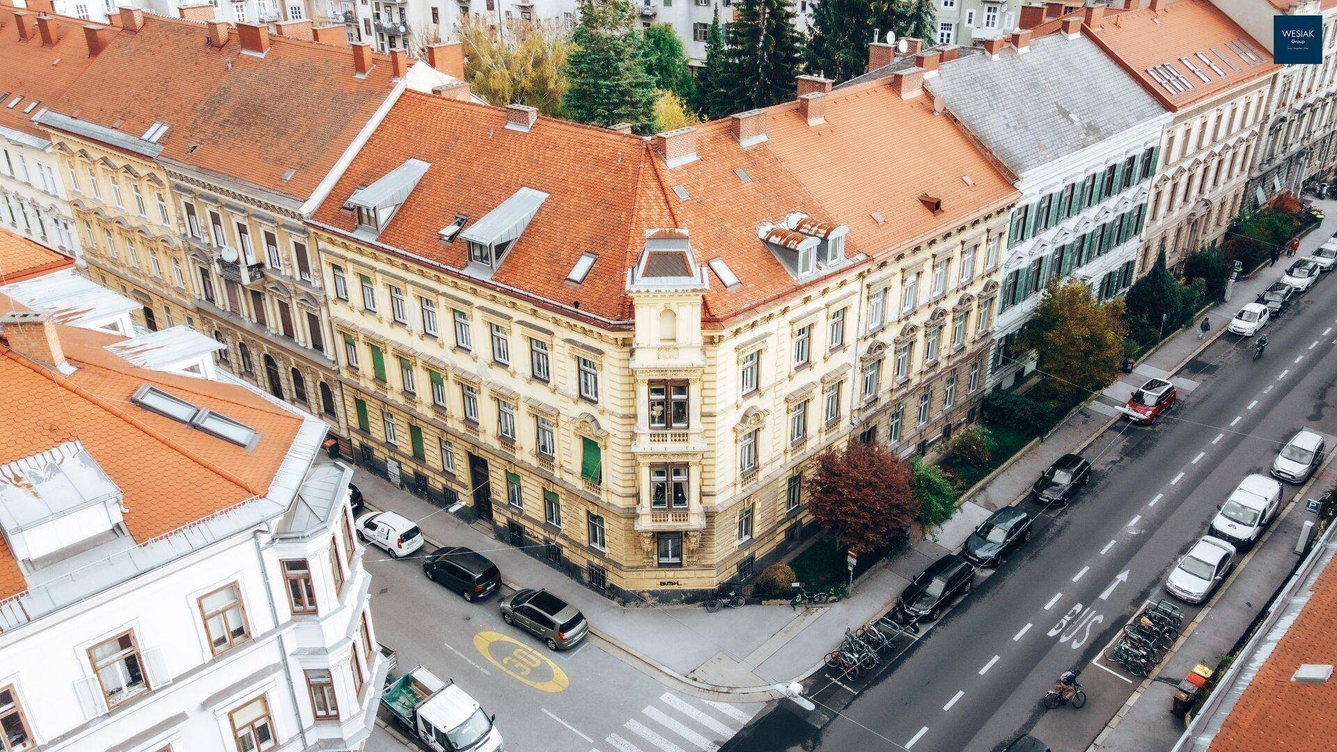 Panoramablick auf das historische Eckgebäude und die belebte Straße mit parkenden Fahrzeugen.