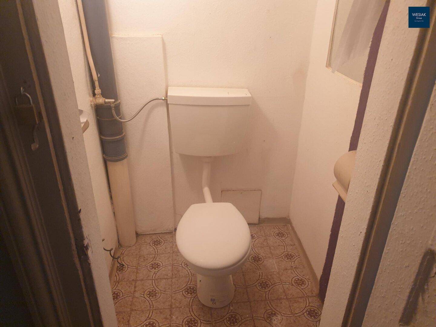 Kleines WC mit einfacher Ausstattung, sichtbaren Rohren und gemustertem Fliesenboden.