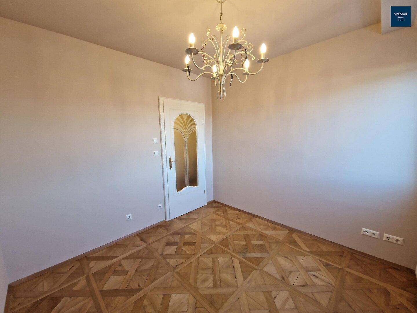 Leerer Raum mit elegantem Parkettboden, weißer Tür und Kronleuchter.