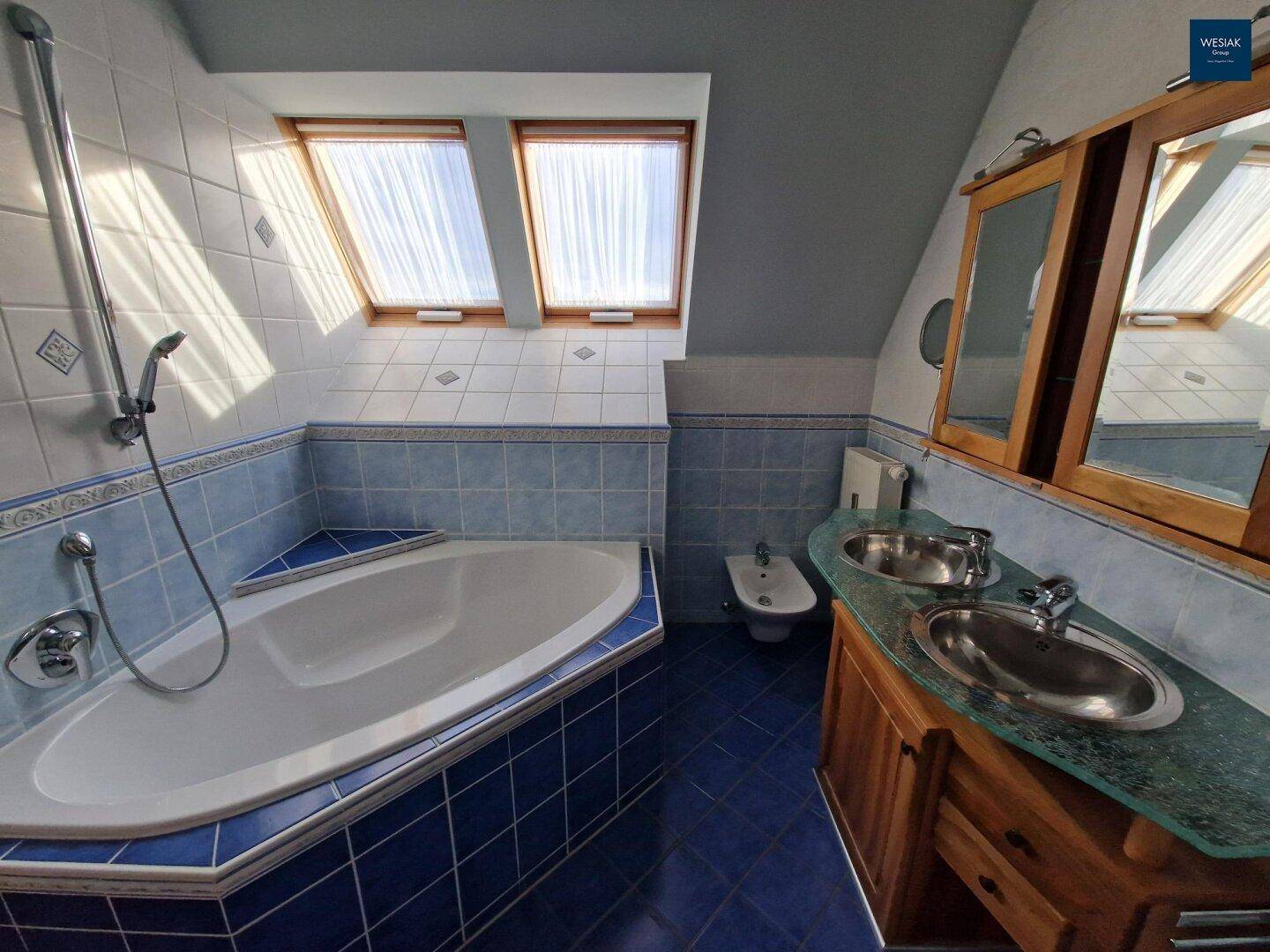 Geräumiges Badezimmer mit blauen Fliesen, Eckbadewanne, Doppelwaschbecken und Dachfenstern.