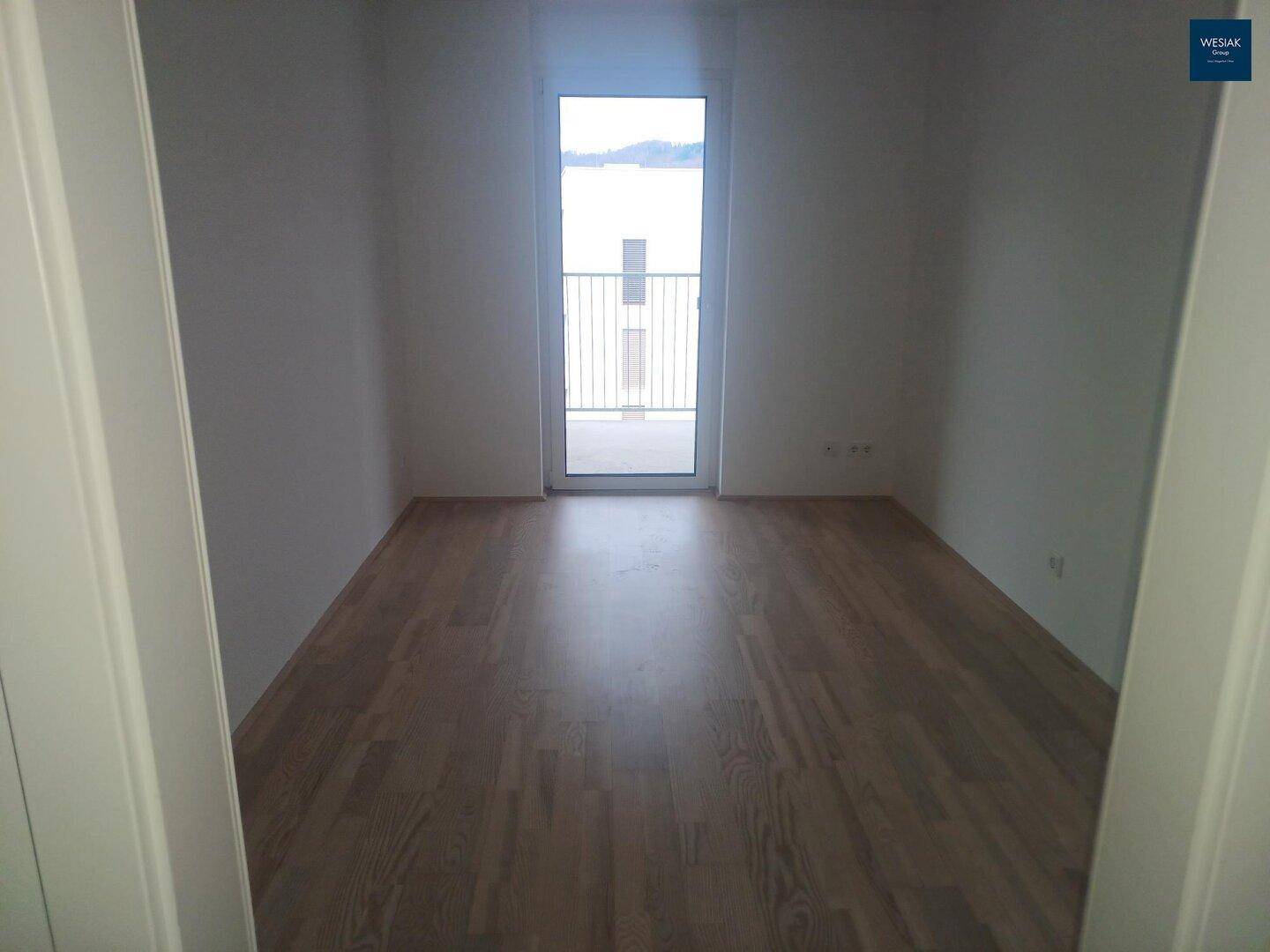 Leeres Zimmer mit Laminatboden und Zugang zu einem kleinen Balkon, ideal als Schlafzimmer.