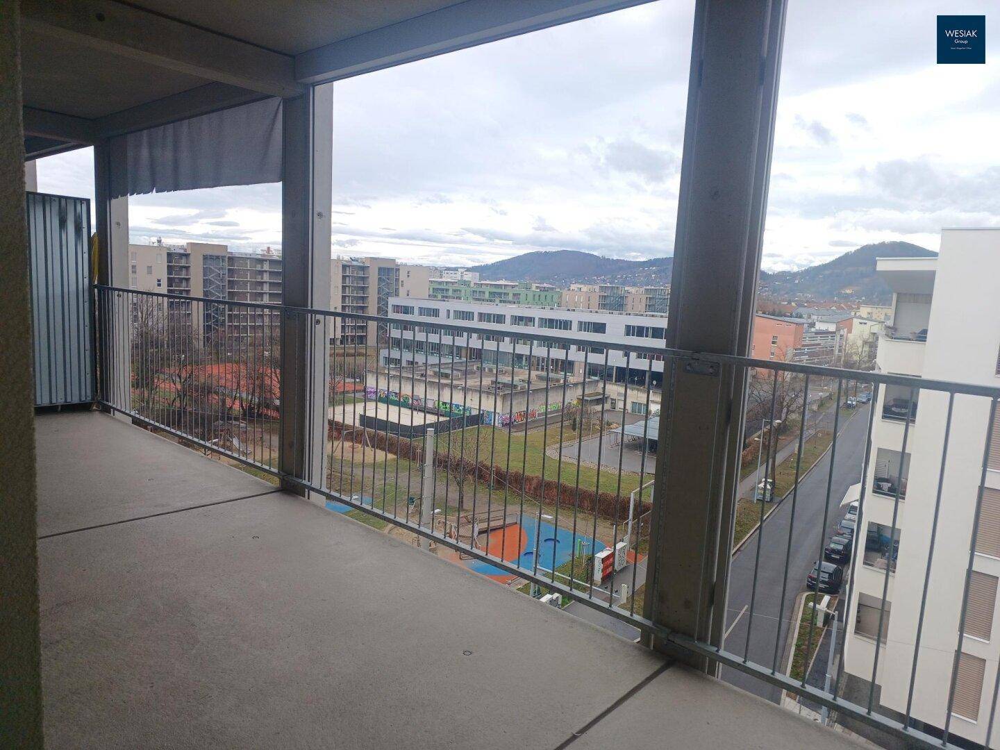 Geräumiger Betonbalkon mit Blick auf die Stadt, Spielplatz und bewaldete Hügel in der Ferne.
