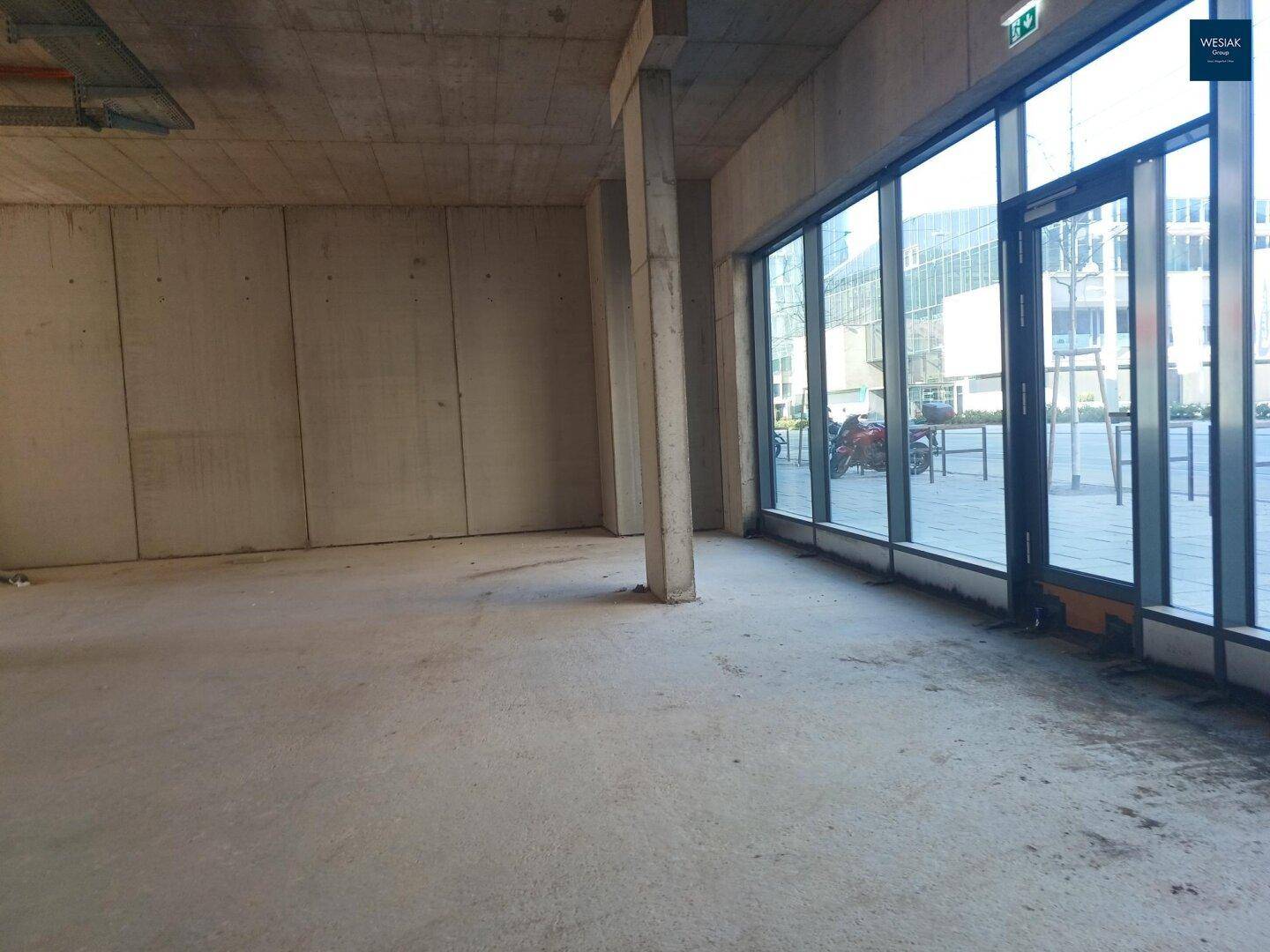 Großer, offener Gewerberaum mit Betonwänden und bodentiefen Fenstern, die viel Tageslicht hereinlassen.
