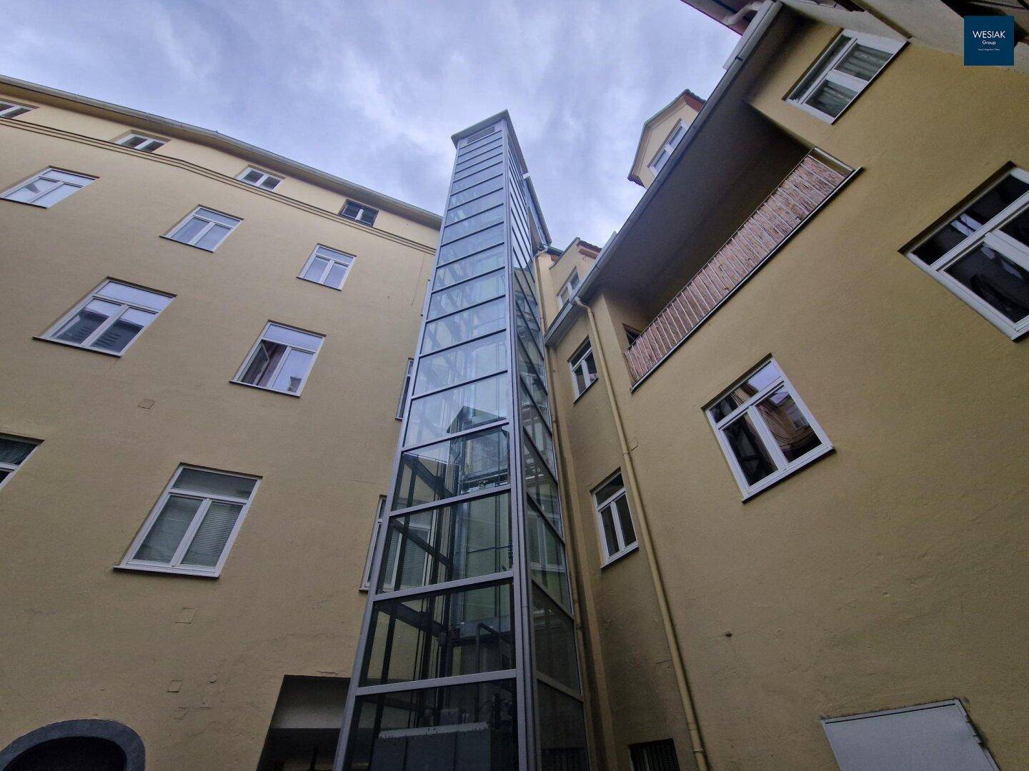 Moderner Glasaufzug an der Fassade eines historischen Gebäudes.