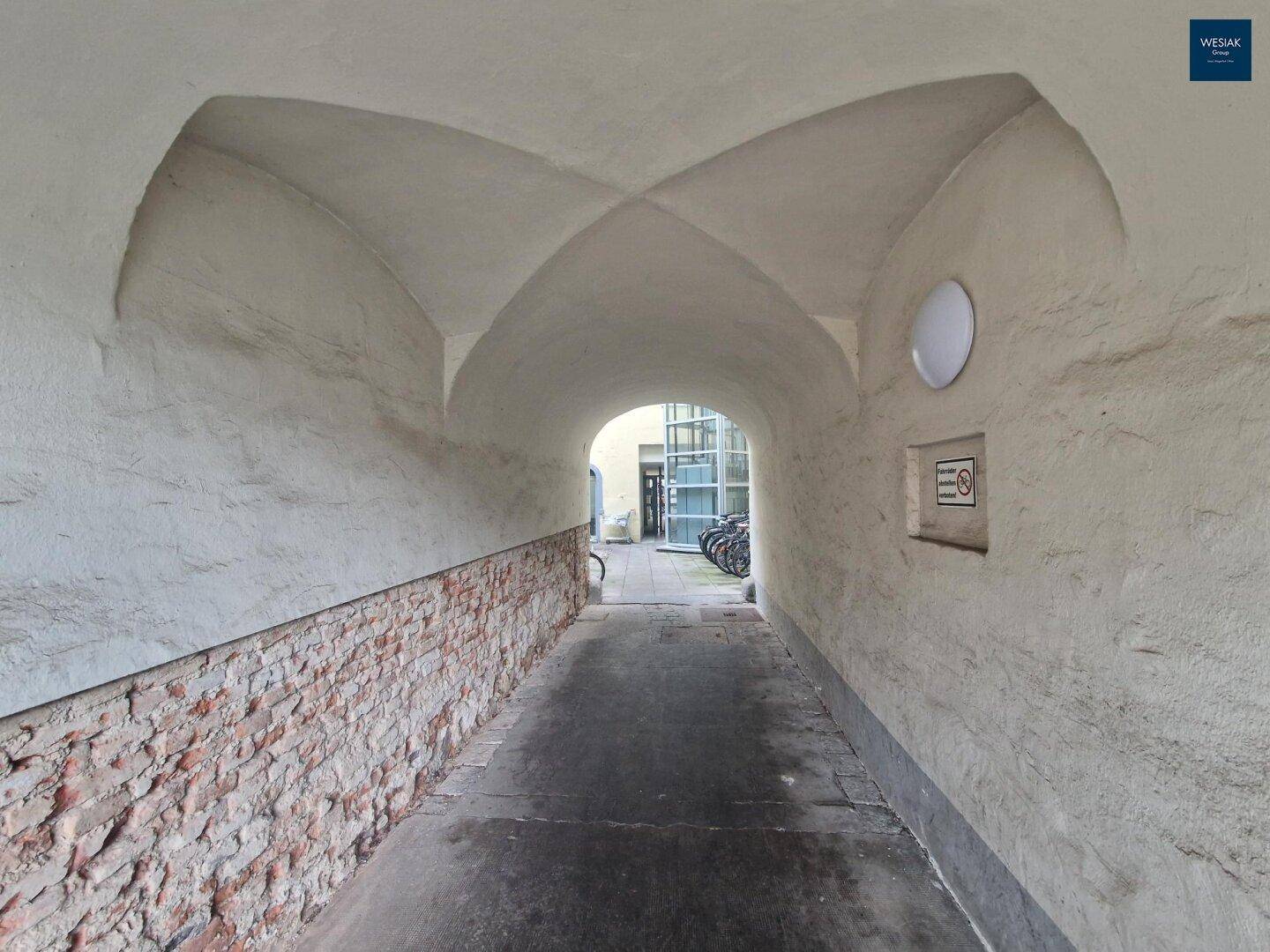 Historischer Torbogen mit Gewölbedecke und Ziegelwand, der zu einem Innenhof führt.