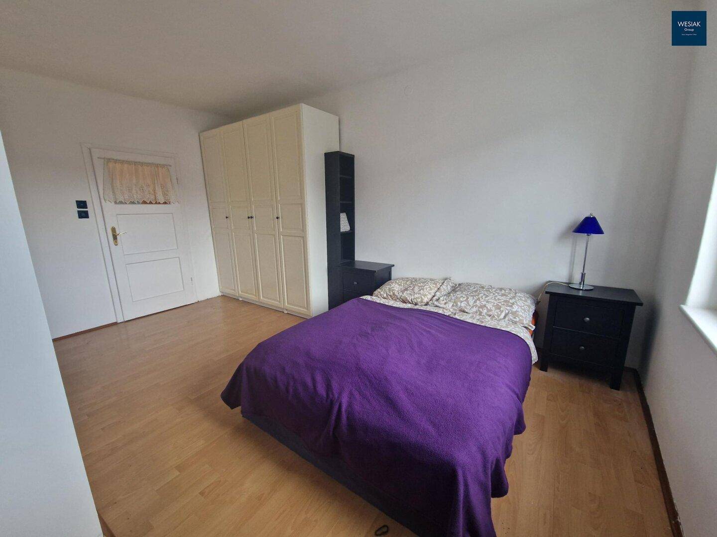 Schlafzimmer mit Holzboden, Kleiderschrank und einem Bett mit violetter Decke.