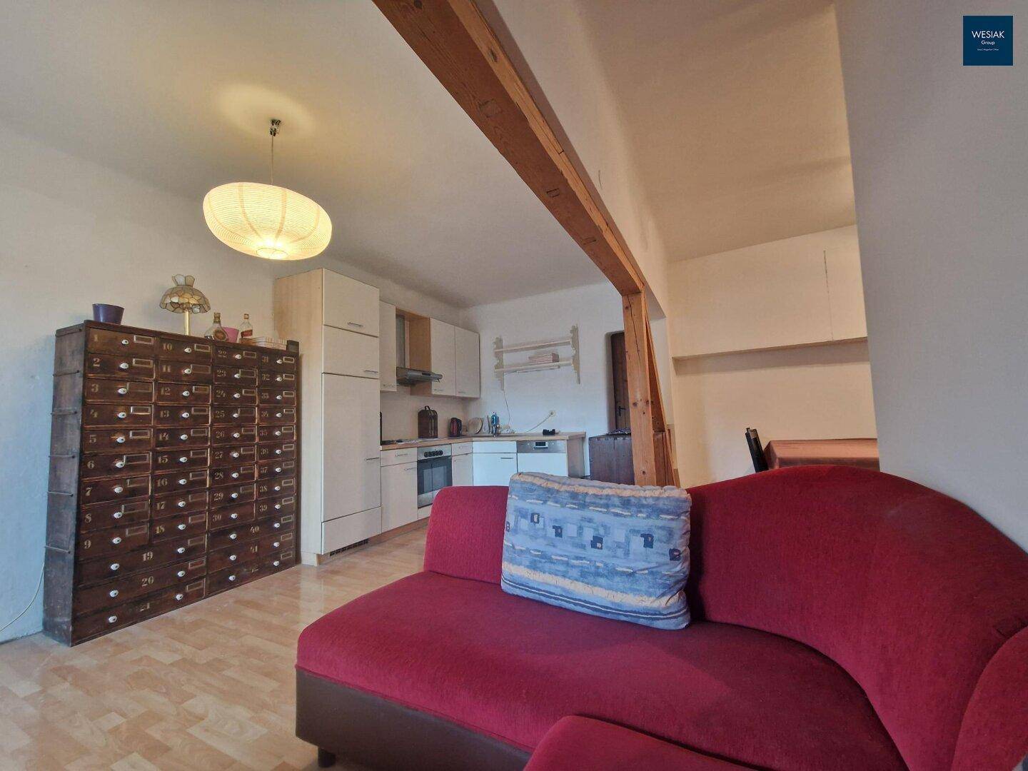 Wohnbereich mit rotem Sofa, Holzbalken und offener Küche im Hintergrund.